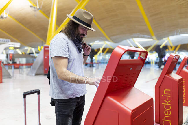 Joven con barba y pelo largo y sombrero comprando billetes de avión en la terminal automática de la aerolínea - foto de stock