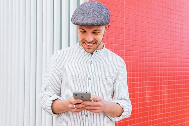 Зміст молодого бородатого хіпстера в повсякденній сорочці поло і кепці перегляду мобільного телефону, стоячи на тлі червоно-білої стіни на сонячному світлі — стокове фото