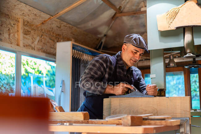 Vista lateral hombre carpintero alisado detalle de madera con gato plano mientras se trabaja en taller de carpintería profesional - foto de stock