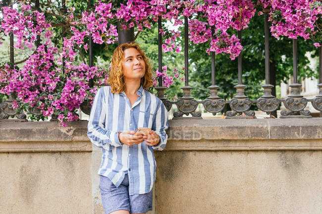 Macho contento con mensajes de pelo largo en el teléfono móvil, mientras que de pie cerca de valla en el parque bajo el árbol en flor con flores de color rosa - foto de stock