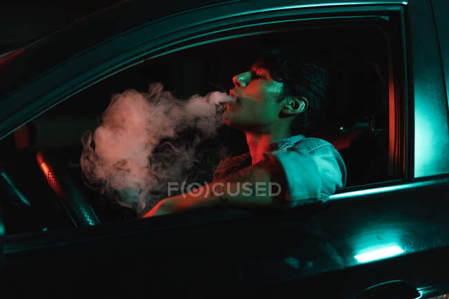 Young latin man sitting in car and smoking under color lights at night — Stock Photo