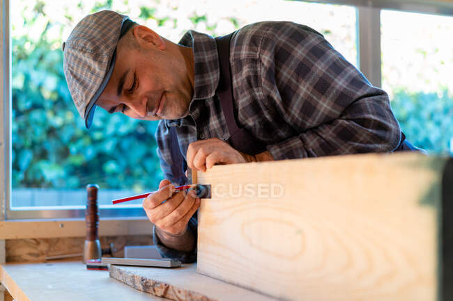 Lenhador profissional positivo com lápis e ferramenta de medição fazendo marcas na placa de madeira enquanto cria objeto de artesanato na oficina de carpintaria — Fotografia de Stock