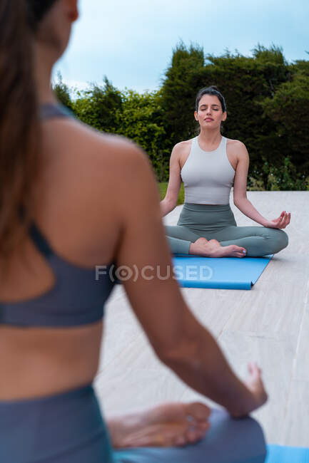 Jovem mindful fêmea com pernas cruzadas e olhos fechados praticando ioga contra a colheita parceiro irreconhecível no pátio — Fotografia de Stock