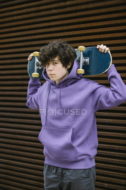 Adolescent sans émotion garçon en vêtements décontractés regardant la caméra tout en se tenant debout avec skateboard derrière la tête sur la rue sur fond flou — Photo de stock