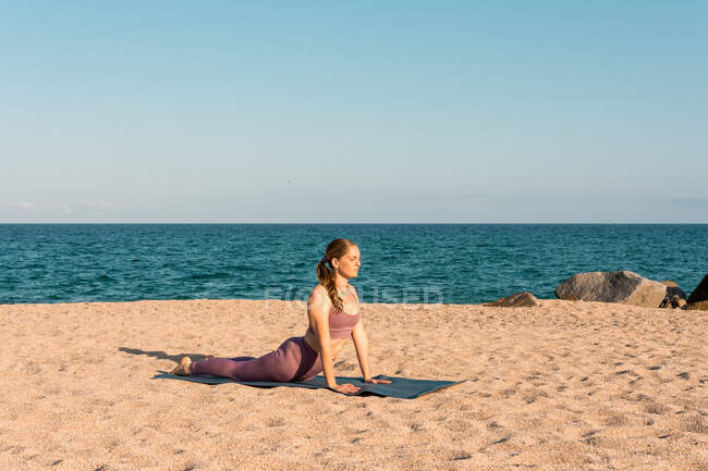 Side view of female doing yoga in Bhujangasana and stretching body on sandy beach on sunny day — Stock Photo