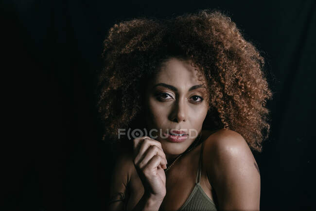 Charmant modèle féminin afro-américain aux cheveux bouclés regardant la caméra en studio sombre — Photo de stock