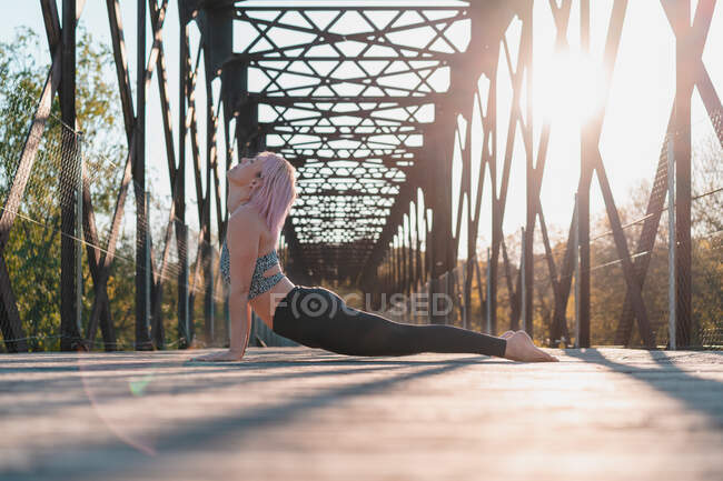 Vista lateral a nivel del suelo de la mujer en ropa deportiva practicando yoga en el sendero del puente en un día soleado - foto de stock