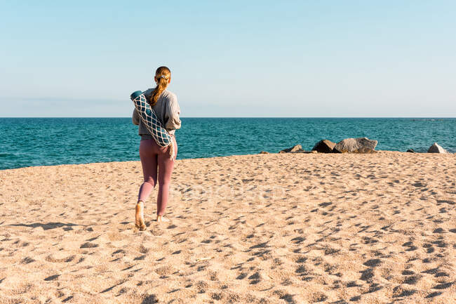 Plein corps vue de dos de méconnaissable de jeune femme pieds nus en vêtements de sport avec tapis de yoga roulé regardant à distance tout en se tenant debout sur la plage de sable près de la mer — Photo de stock