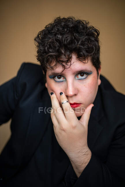 Homme androgyne immobile avec lèvres rouges et fard à paupières lumineux avec la main sur le visage et regardant la caméra — Photo de stock