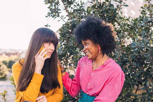 Черная улыбающаяся лесбиянка смотрит на девушку, сидящую в парке и разговаривающую по смартфону в выходные — стоковое фото