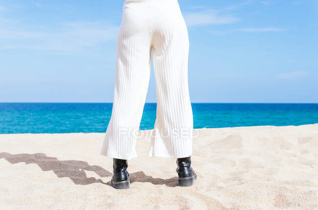 Rückansicht einer anonymen Frau in Stiefeln, die an einem sonnigen Tag am Sandstrand in Richtung ruhiges blaues Meer steht — Stockfoto