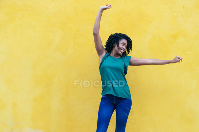 Смеющаяся молодая женщина перед желтой стеной с поднятыми руками — стоковое фото