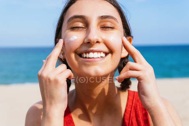 Delighted female smearing sunscreen lotion on face while chilling on seashore on sunny day in summer — Stock Photo