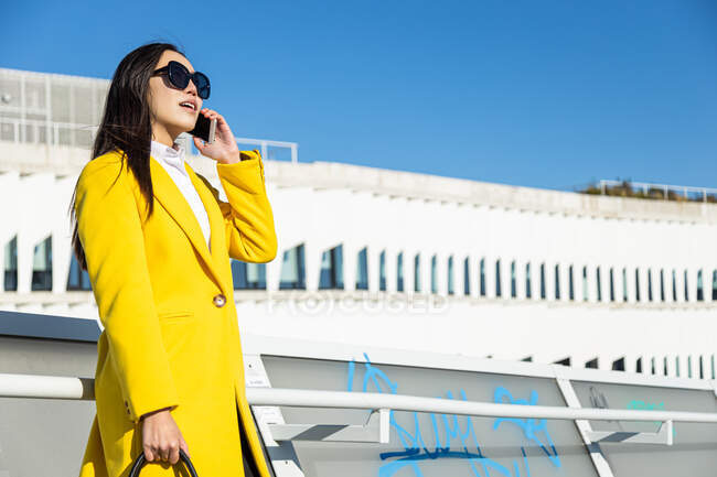 Mujer de negocios asiática sonriente con abrigo amarillo y teléfono inteligente caminando en la calle con el edificio en el fondo - foto de stock