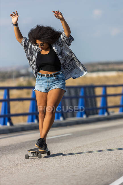 Young and afro woman skating long board by an empty bridge at sunset, front view — Stock Photo