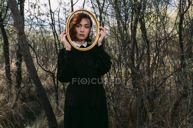 Mujer sin emociones en vestido negro de pie en el bosque de otoño en Halloween y mirando a la cámara a través del agujero en el marco de madera - foto de stock