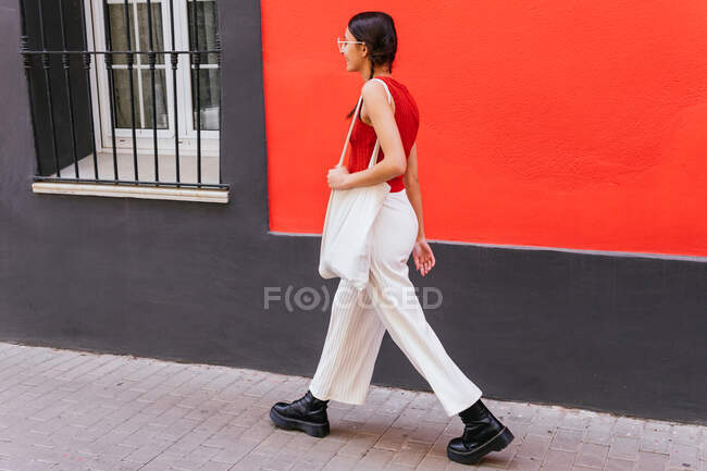 Vista lateral del contenido femenino de moda con bolsa textil ecológica caminando por la calle cerca del edificio rojo en la ciudad - foto de stock