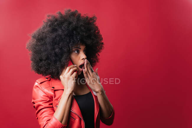 Amazed African American female speaking on mobile phone and covering mouth while listening to rumors on red background in studio — Stock Photo