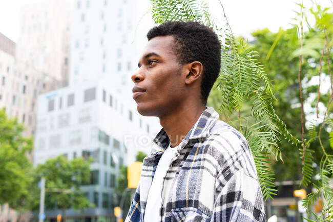 Vista lateral do homem afro-americano elegante e pouco emocional em pé na cidade olhando para longe — Fotografia de Stock