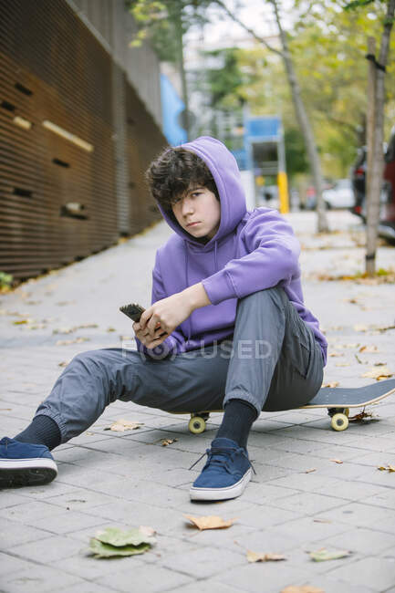 Seriöser männlicher Teenager in Freizeitkleidung sitzt auf einem Skateboard und schaut in die Kamera, während er auf gepflastertem Bürgersteig in der Stadt mit dem Handy surft — Stockfoto
