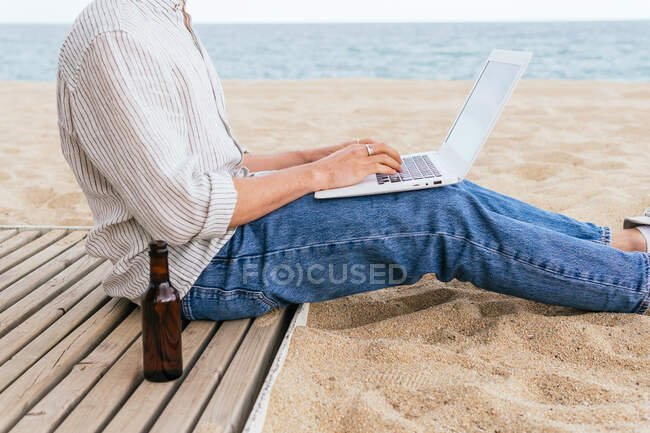 Side view of cropped unrecognizable male blogger in stylish wear and hat sitting with bottle of beer on sandy beach and typing on laptop during summer holidays on seashore — Stock Photo