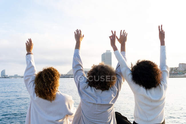 Back view of anonymous friends with curly hair sitting close with arms raised against cityscape and embankment in sunlight — Stock Photo