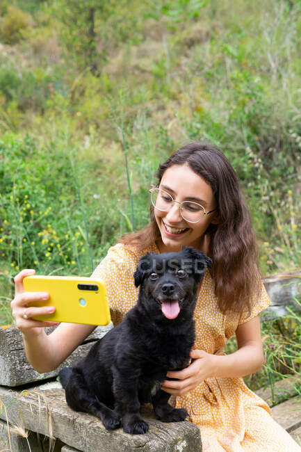 Alegre hembra tomando uno mismo tiro con negro peludo cachorro mientras se utiliza el teléfono inteligente y sentado en un banco de madera en el campo - foto de stock