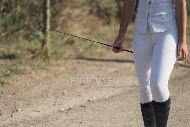 Cortar sem rosto feminino equestre em uniforme e com chicote em pé na arena arenosa no clube equino — Fotografia de Stock
