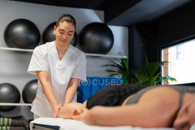 Joyeuse masseuse en peignoir blanc massant le mollet d'une patiente des cultures lors d'une séance de physiothérapie en clinique — Photo de stock