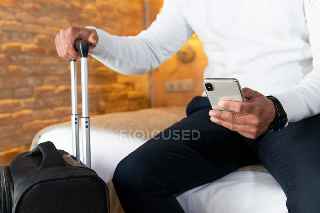 Männlicher Reisender sitzt auf Bett in der Nähe von Koffer und surft Handy im Hotelzimmer — Stockfoto