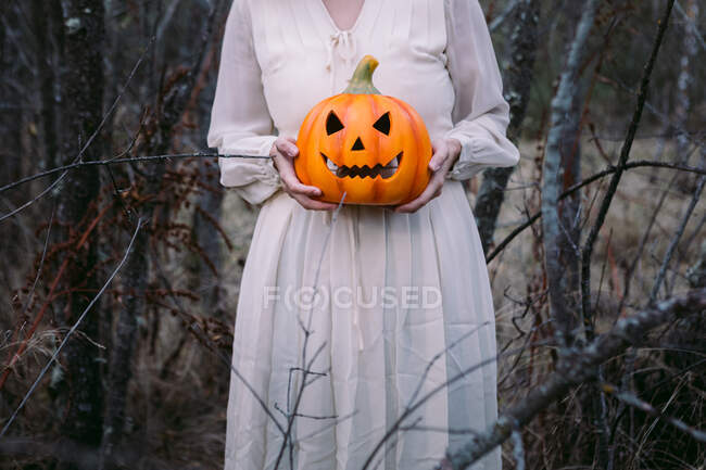 Femme en robe noire debout avec lanterne de citrouille éclatante dans les bois sombres sur Halloween et en regardant la caméra — Photo de stock