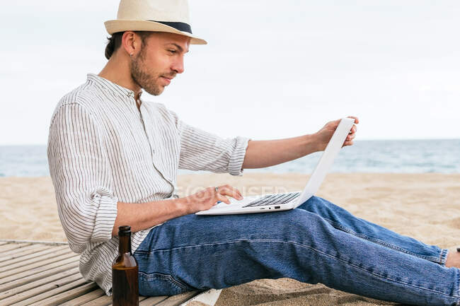 Вид збоку молодого чоловіка блогер в стильному одязі і капелюсі сидить з пляшкою пива на піщаному пляжі і друкує на ноутбуці під час літніх канікул на березі моря — стокове фото