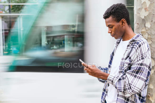 Vista lateral de mensajería masculina afroamericana segura en el teléfono móvil mientras está de pie en la calle de la ciudad - foto de stock