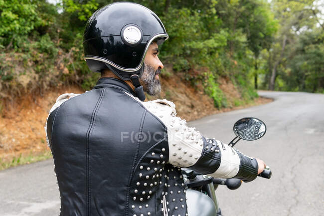 Вид сзади бородатый этнический байкер в черной кожаной куртке и шлеме верхом на современном мотоцикле по асфальтированной дороге среди пышных зеленых деревьев, растущих в горной долине — стоковое фото
