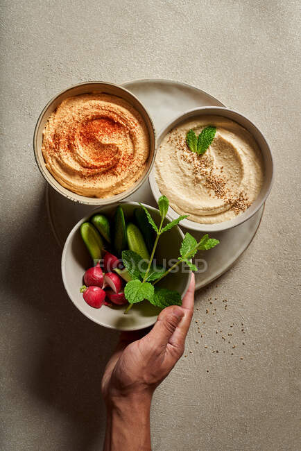 Top view of crop person holding bowls with assorted hummus served on table with fresh cucumbers and radish — Stock Photo