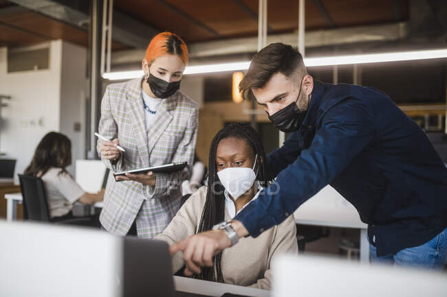 Group of diverse coworkers in protective masks gathering at table with laptop and discussing project while working together in contemporary workspace — Stock Photo