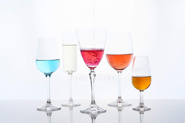 Разнообразные разноцветные коктейли в хрустальных бокалах различной формы, подаваемые на зеркальном столе на белом фоне — стоковое фото