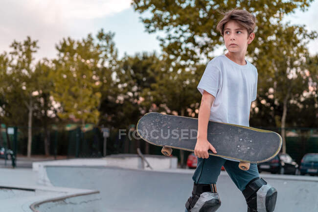 Teenage boy in protective gear standing with skateboard in skate park and looking away — Stock Photo