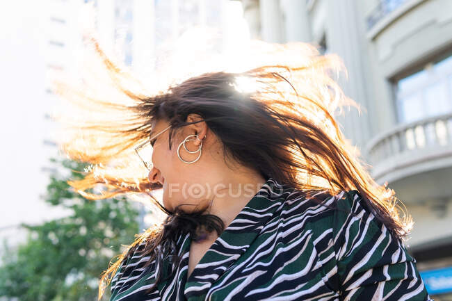 Bajo ángulo de alegre joven hembra en traje elegante y pendientes sacudiendo el cabello a la luz del sol en la calle urbana - foto de stock