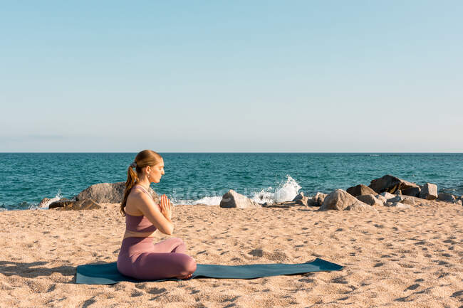 Seitenansicht einer gelassenen Frau, die in Padmasana sitzt und Yoga macht, während sie mit geschlossenen Augen am sandigen Meeresufer meditiert — Stockfoto