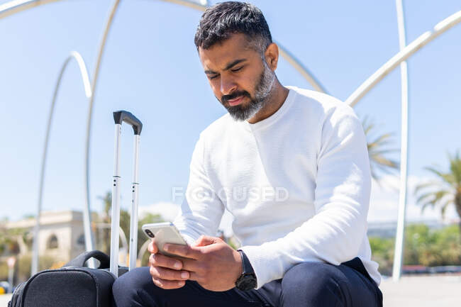 Hombre adulto concentrado en ropa casual inteligente que navega por el teléfono móvil y se sienta en la calle soleada con la maleta - foto de stock