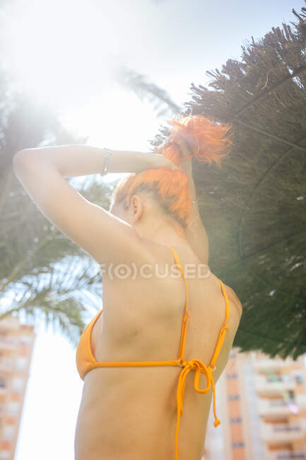 Задний вид анонимной молодой рыжей женщины, делающей хвостик на пляже в солнечный день летом — стоковое фото