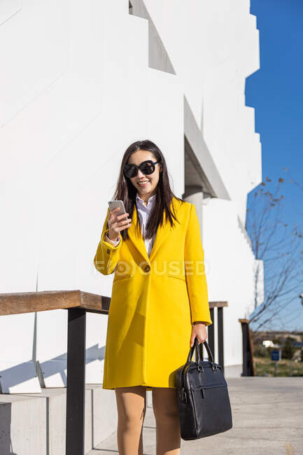 Souriant asiatique femme d'affaires avec manteau jaune et téléphone intelligent marche dans la rue avec bâtiment en arrière-plan — Photo de stock