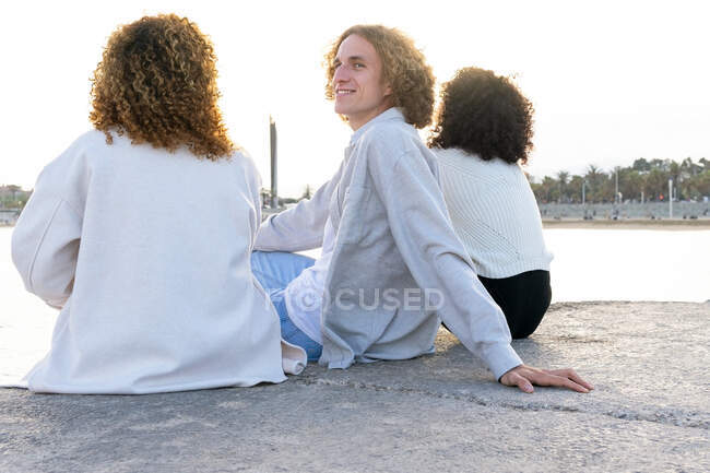 Улыбающийся мужчина отдыхает с подругами на городской набережной и оглядывается через плечо — стоковое фото