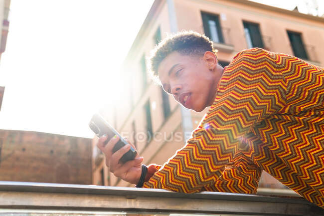 De baixo vista lateral do jovem hispânico cara com cabelo afro em elegante roupa colorida navegando telefone celular, enquanto se inclina em trilhos perto do edifício urbano sob a luz do sol — Fotografia de Stock