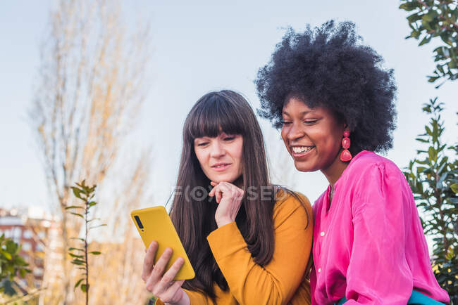 Feliz casal multiétnico de mulheres homossexuais sentadas na rua e navegando telefone celular juntos — Fotografia de Stock