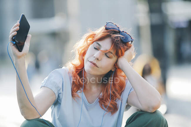 Femme rêveuse avec les cheveux roux assis dans la rue et écoutant de la musique dans les écouteurs tout en appréciant les chansons avec les yeux fermés et les bras tendus — Photo de stock