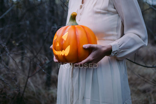 Cropped unrecognizable female in white dress standing with glowing pumpkin lantern in the woods on Halloween and looking away — Stock Photo