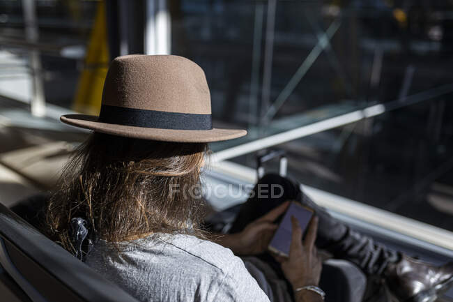 O cara de chapéu no aeroporto na sala de espera sentado esperando por seu voo, com fones de ouvido sem fio para ouvir música enquanto conversa com seu telefone inteligente, visão traseira — Fotografia de Stock