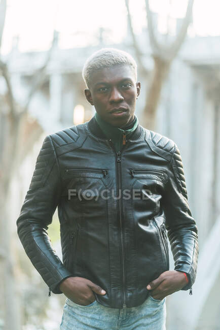 Confident African American male wearing trendy leather jacket standing in autumn park and looking at camera — Stock Photo